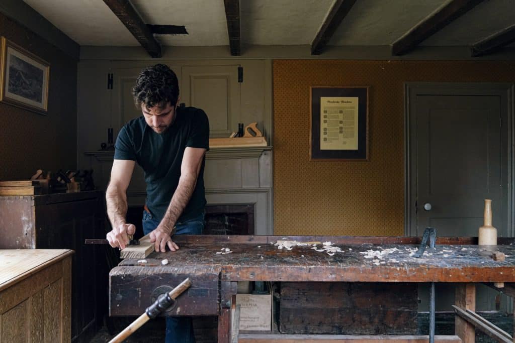 A man in a black t-shirt works on a wooden piece at a rustic workbench in a carpentry-styled room.