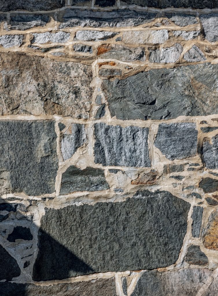 Close-up of a stone wall with irregularly shaped, rough-textured stones in various shades of gray and beige, with mortar filling the gaps.