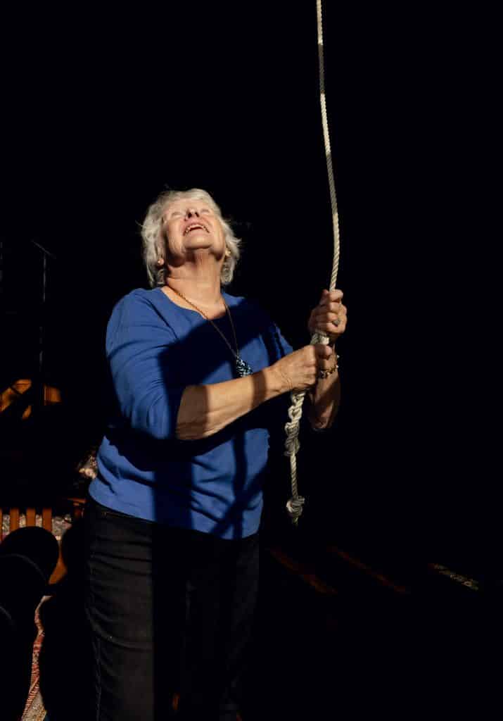 Person with white hair in a blue shirt pulling on a rope, looking upward, against a dark background.