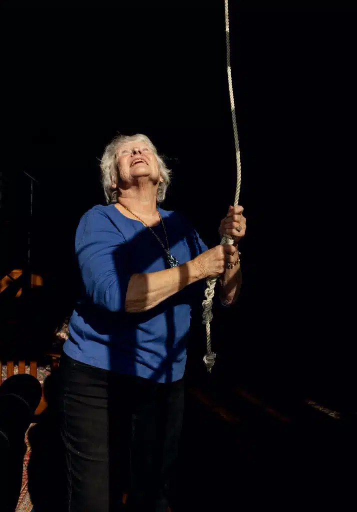 Person with white hair in a blue shirt pulling on a rope, looking upward, against a dark background.