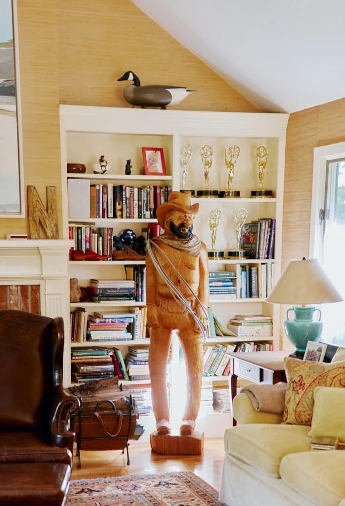 Room with a wooden cowboy statue, bookshelf filled with books and awards, a duck figurine, a leather chair, and a side table with a lamp.