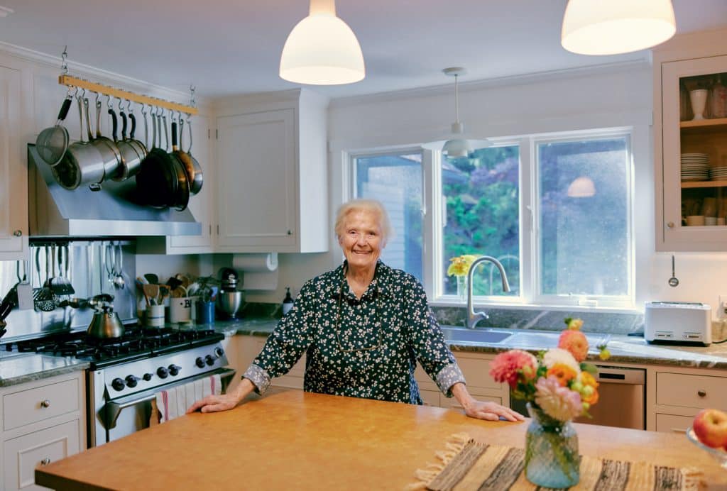 An elderly person stands in a kitchen with hanging pots and a flower vase on the counter.