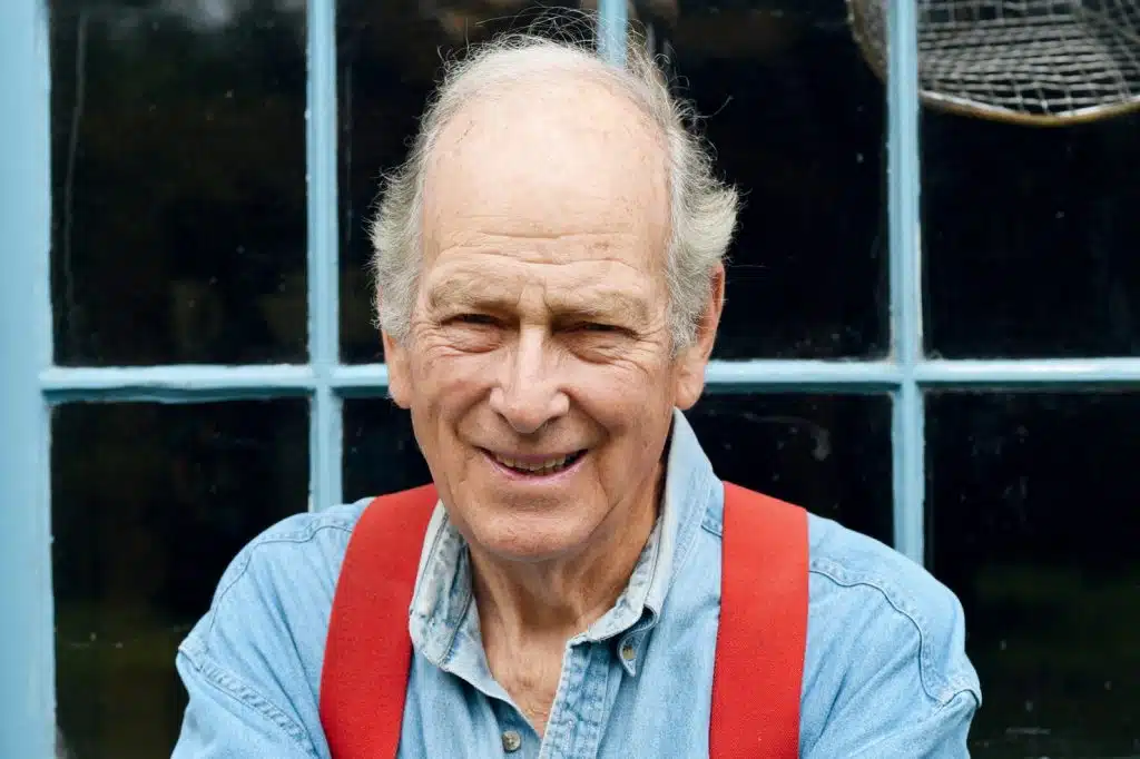 Elderly man smiling, wearing a blue shirt and red suspenders, standing in front of a window.