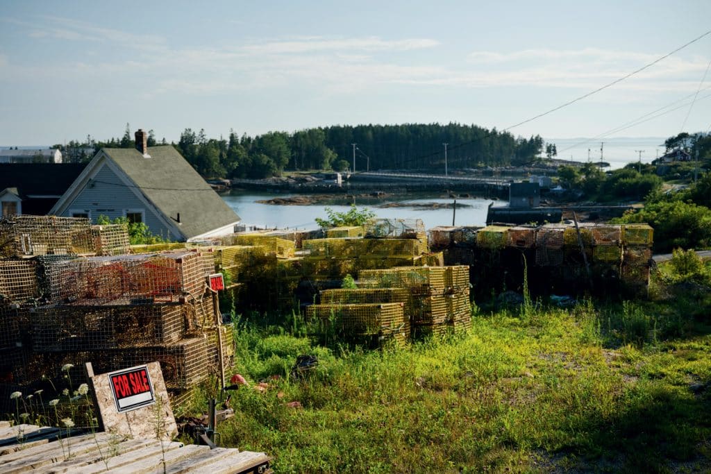 Stacks of lobster traps on grass with a "For Sale" sign in front. A house and trees are in the background, near a body of water under a cloudy sky.