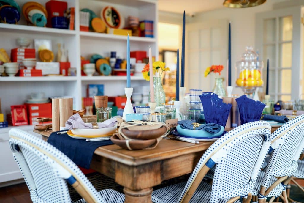 A dining table set with plates, candles, and glassware. Blue and white chairs surround the table. Shelves in the background hold various colorful dishes and items.