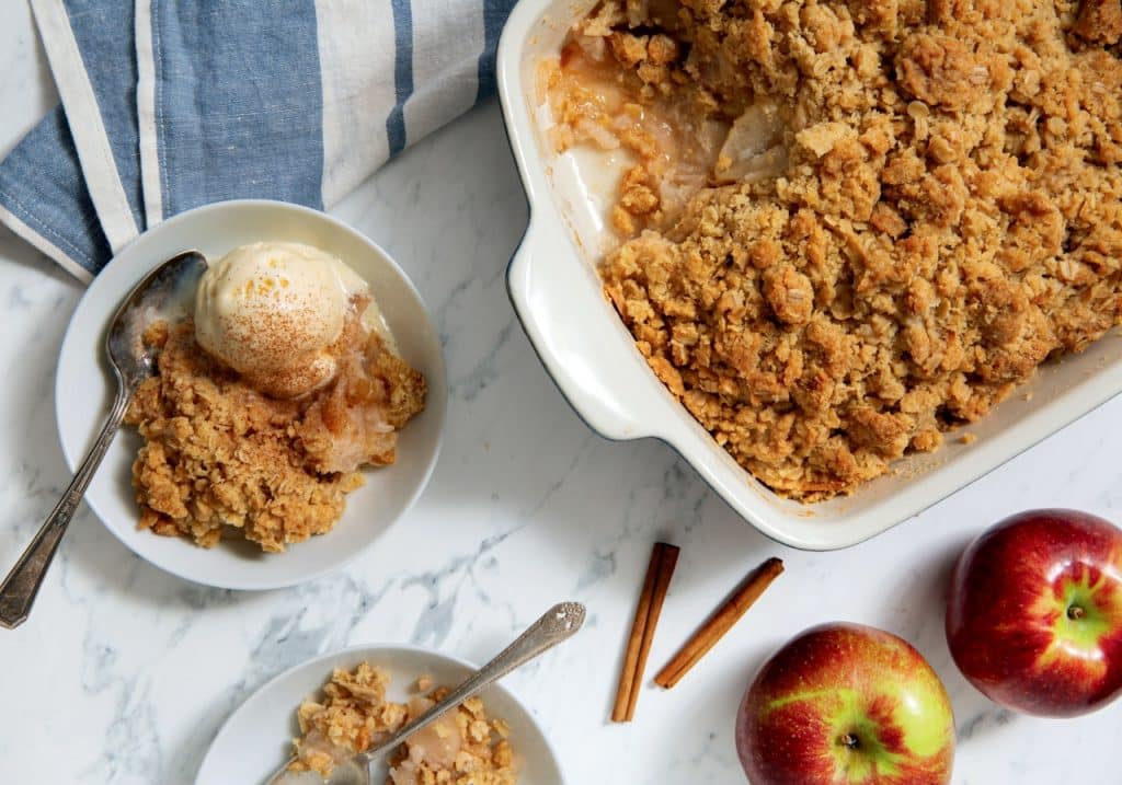 Apple crumble in a baking dish with a serving on a plate, topped with ice cream. Two whole apples, cinnamon sticks, and a striped cloth are nearby on a marble surface.