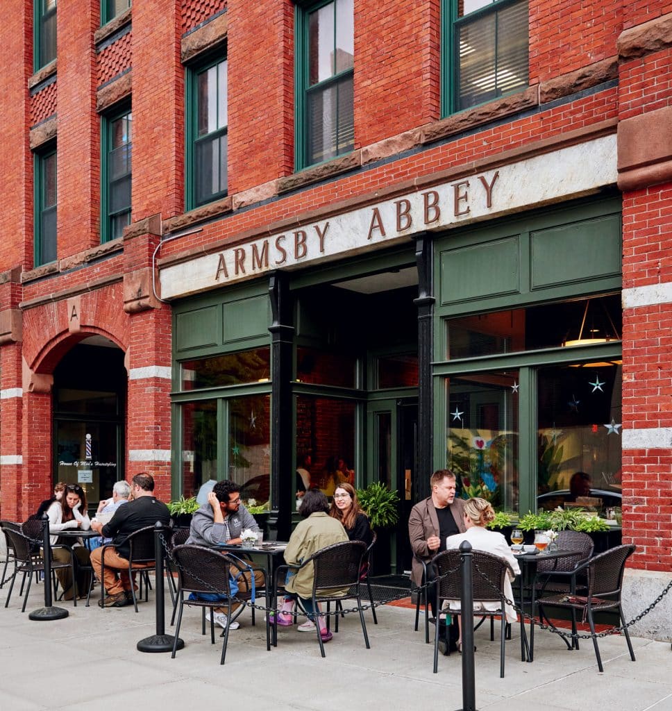 People sitting at tables enjoying food and drinks outside a brick building with a sign reading "Armsby Abbey.