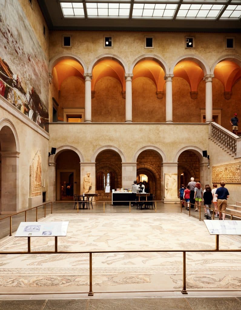 A museum interior with arches, columns, and a large mural. People are walking and observing exhibits, including a mosaic on the floor.