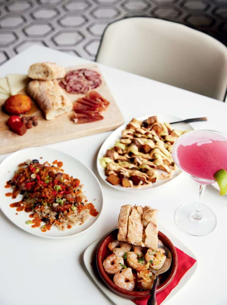 A table with various dishes including shrimp in a bowl, a charcuterie board, a plate with toppings, a dish with drizzled sauce, and a pink cocktail garnished with a lime.