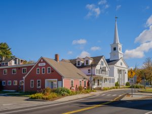 A white church with a tall steeple sits next to a red building on a sunny day. A road sign indicates a turn to the right.