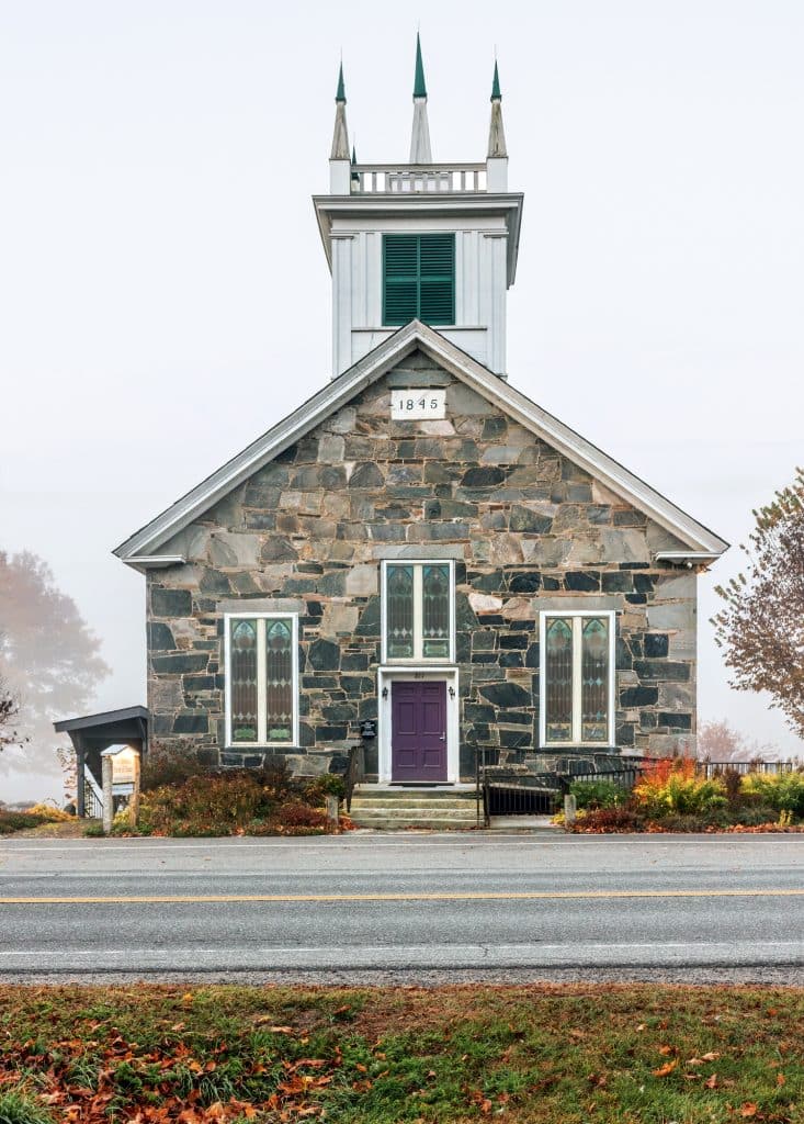 A stone church built in 1845 with a purple door, tall windows, and a tower featuring green-capped spires stands by a road, surrounded by autumn foliage.