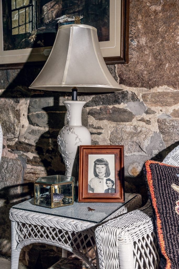 White wicker table with a lamp, framed photo, glass box, and crochet blanket, set against a stone wall.