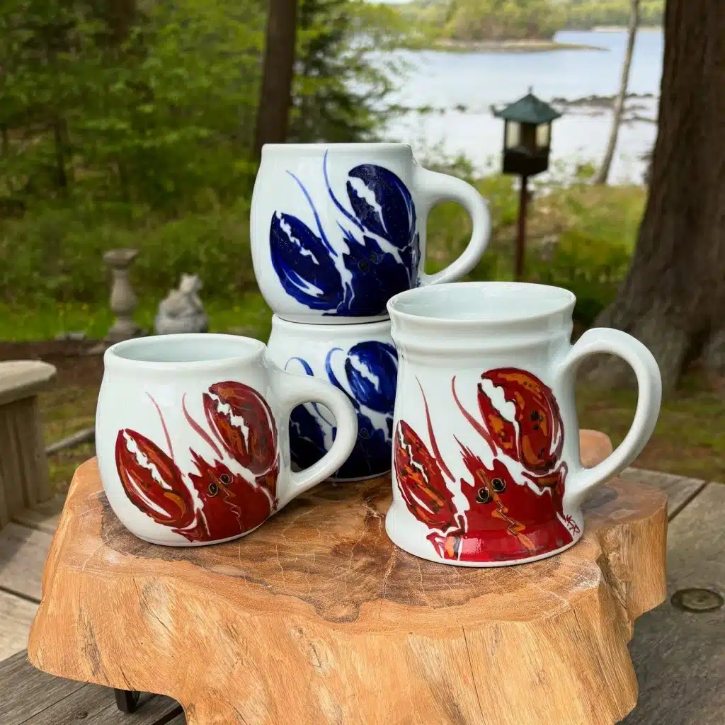 New England pottery. Four ceramic mugs with blue and red lobster designs sit on a wooden stump outdoors near trees and water.