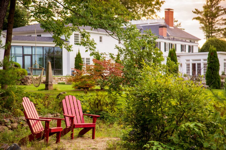 Two red Adirondack chairs are set on a stone patio amid greenery, facing a large white house with multiple windows and a sunroom in the background.