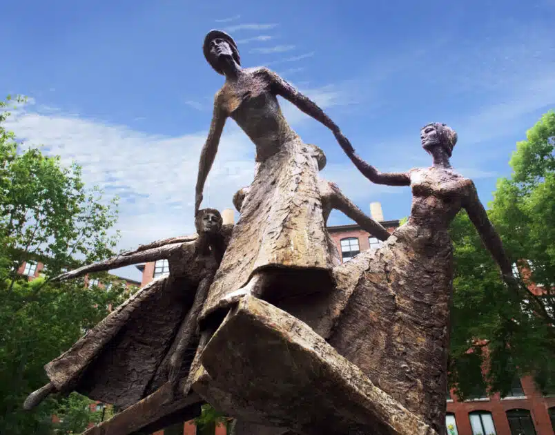 Bronze sculpture of four women holding hands in a circle, outdoors, with trees and brick buildings in the background under a blue sky.