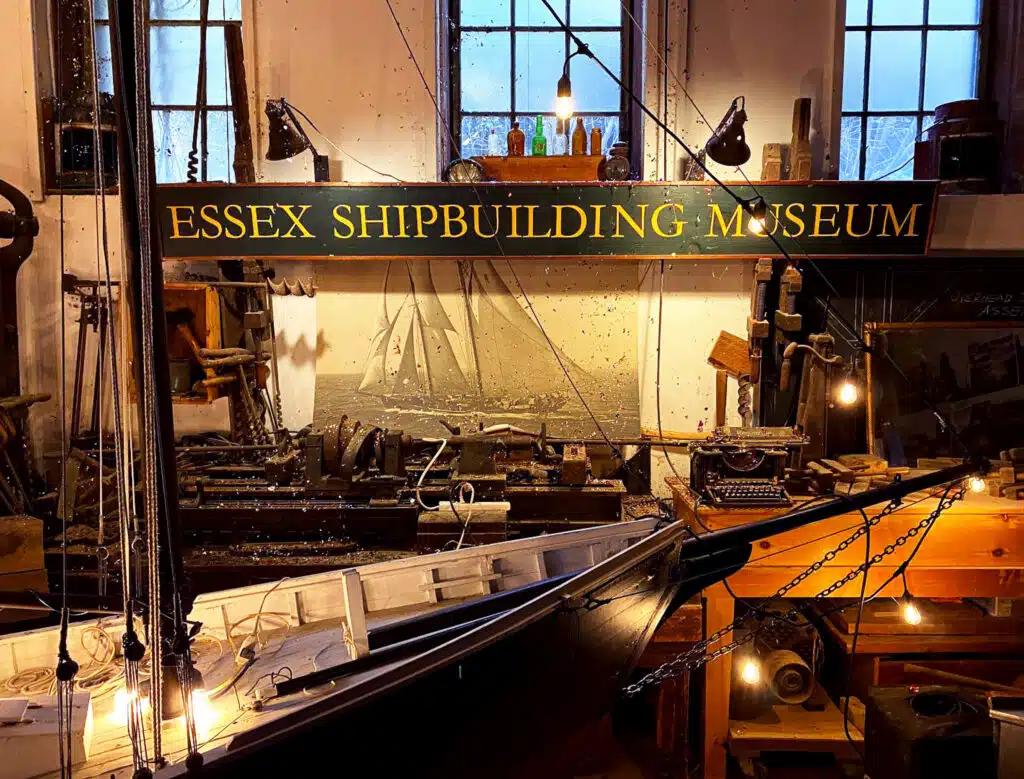 A model ship and antique shipbuilding tools displayed inside the Essex Shipbuilding Museum, with a green and gold sign above.