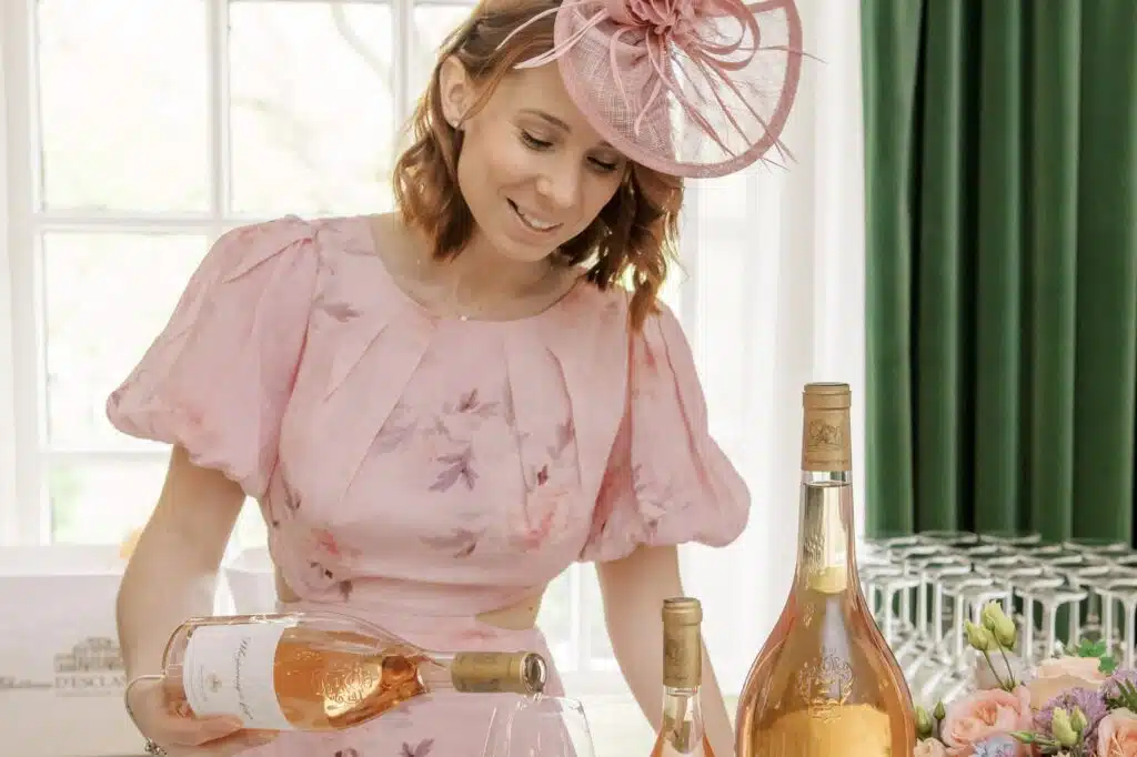 Woman in a pink dress and fascinator pouring rosé wine at a table with wine bottles and glasses.