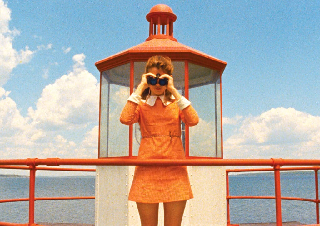 A person in an orange dress stands on a lighthouse platform, looking through binoculars toward the camera under a blue sky with clouds.