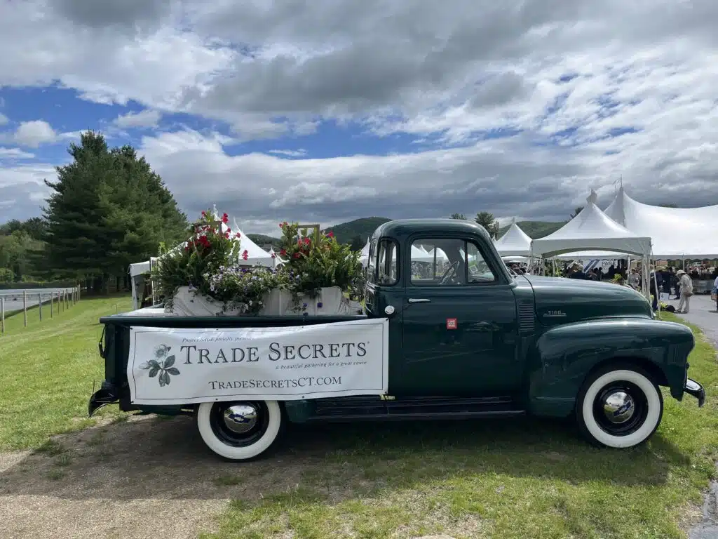 A vintage green truck with flowers in the bed and a "Trade Secrets" banner parked on grass by white tents.