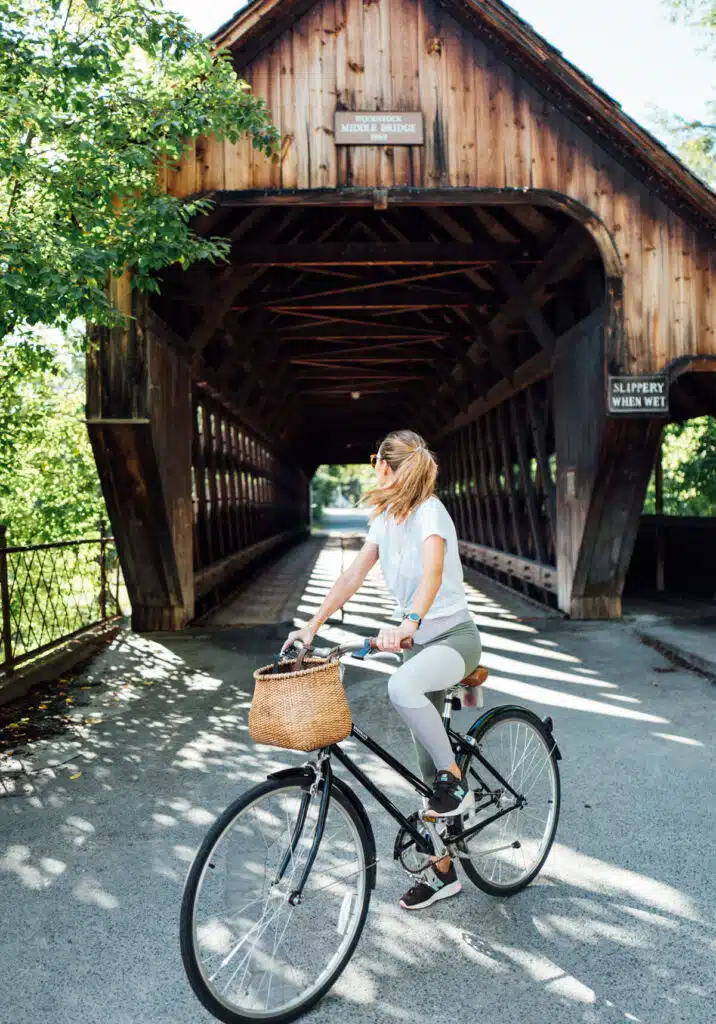 Jess Kirby. A woman rides a bicycle with a basket in front of a wooden covered bridge on a sunny day.