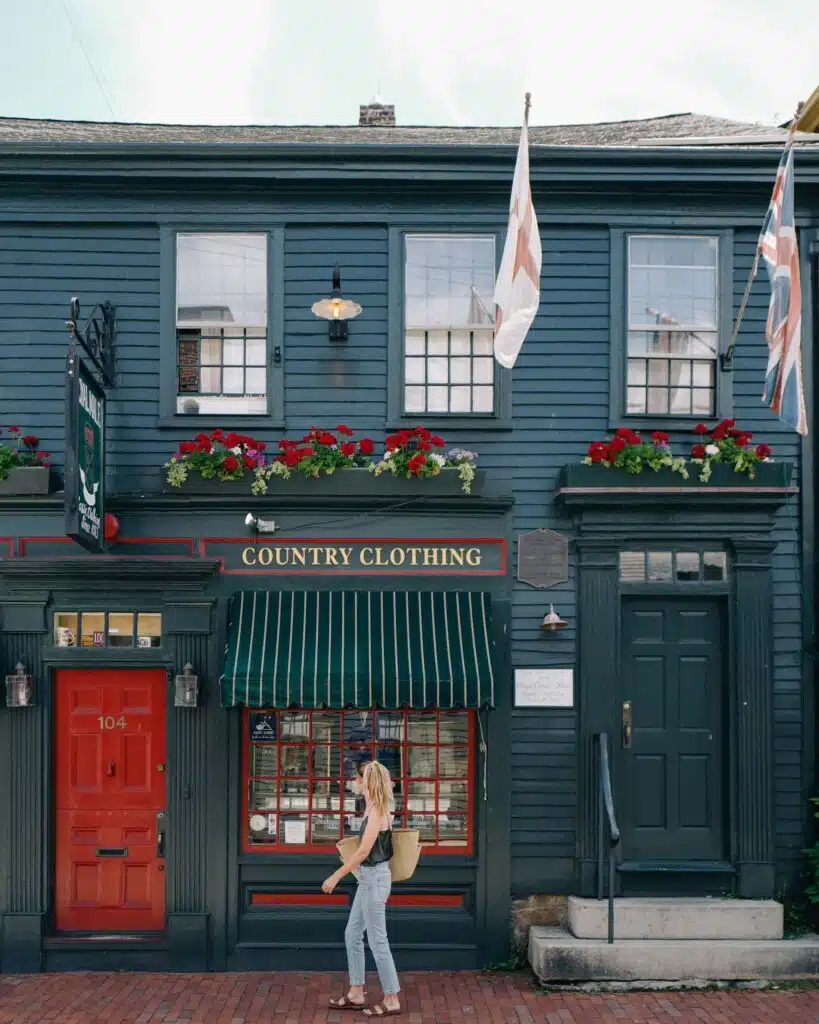 A woman walks past a blue two-story building with flower boxes, striped awning, and a sign that reads "Country Clothing." Flags are displayed on either side of the entrance.