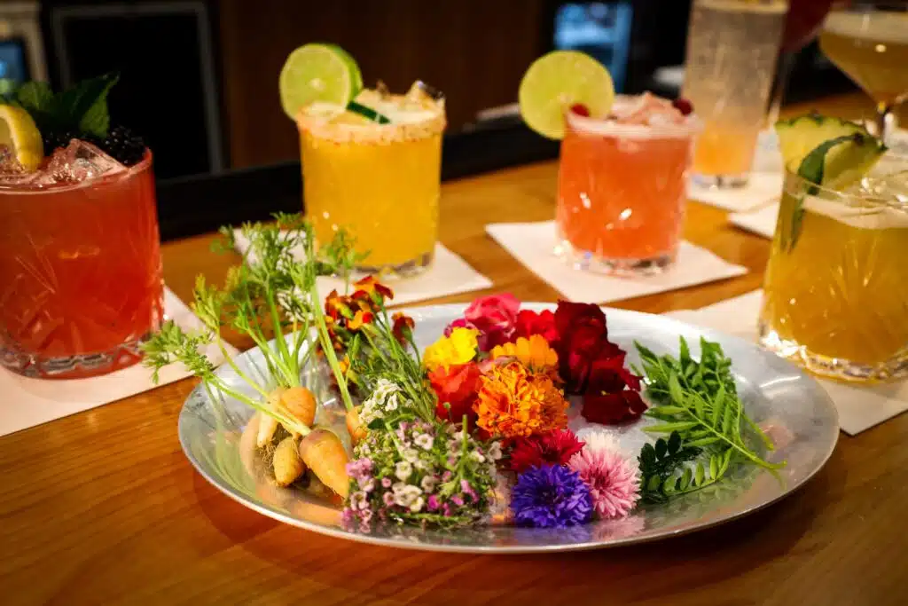 Colorful cocktails with ice and lime on a bar, next to a plate of fresh edible flowers and vegetables.