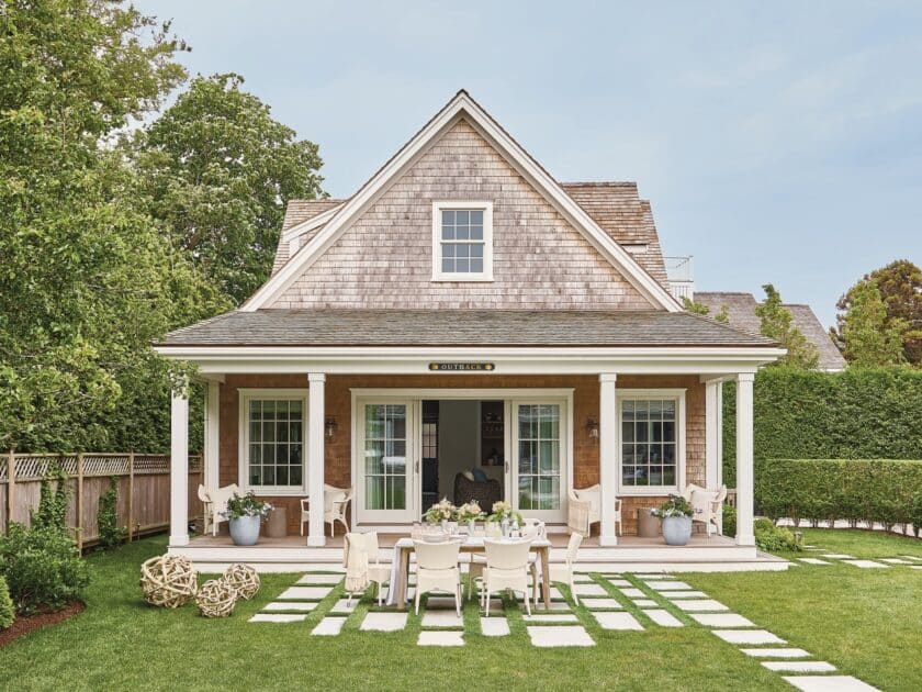 Coastal Charm Behind the Hedge: A Featured Cottage from "Nantucket Looms: A Legacy of Style" - Cozy house with wooden shingles, columns, and a front porch. A dining table with chairs is set on the lawn, surrounded by hedges and a tree.