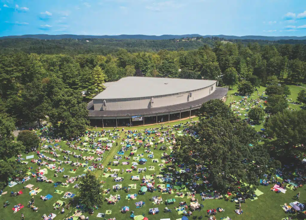 Aerial view of a large outdoor amphitheater surrounded by trees, with crowds of people sitting on blankets and lawn chairs spread across the grassy area.