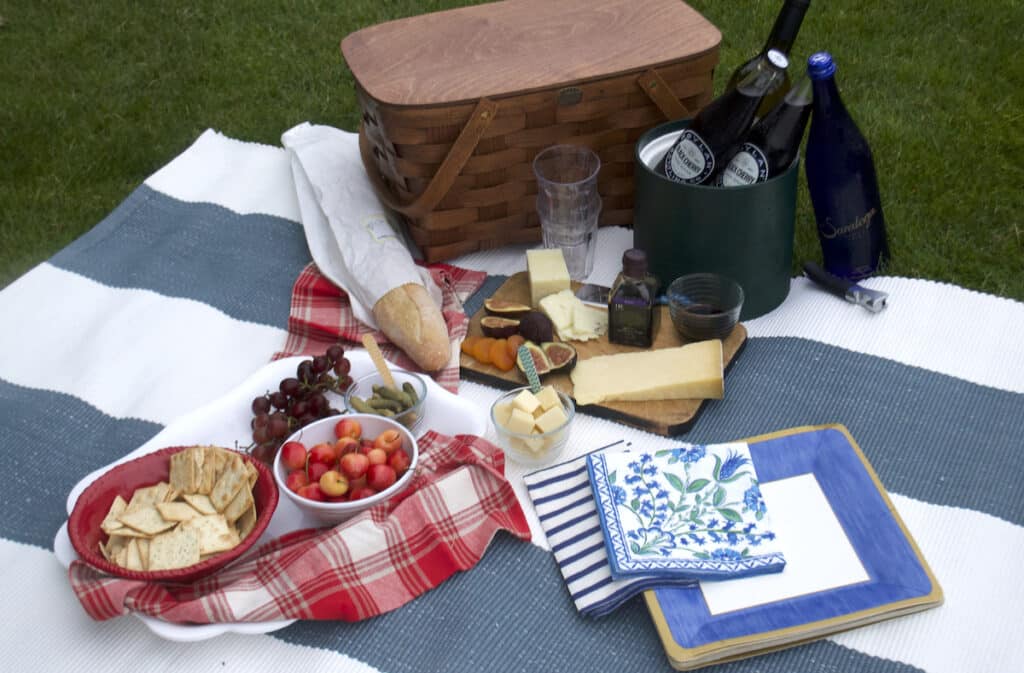 A picnic setup with a woven basket, bread, cheese, fruit, crackers, olives, bottled drinks, plates, napkins, and glasses arranged on a blanket on the grass.