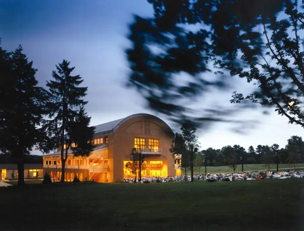 A warmly-lit concert hall with large windows stands among trees at dusk, with a crowd seated outside on the lawn.