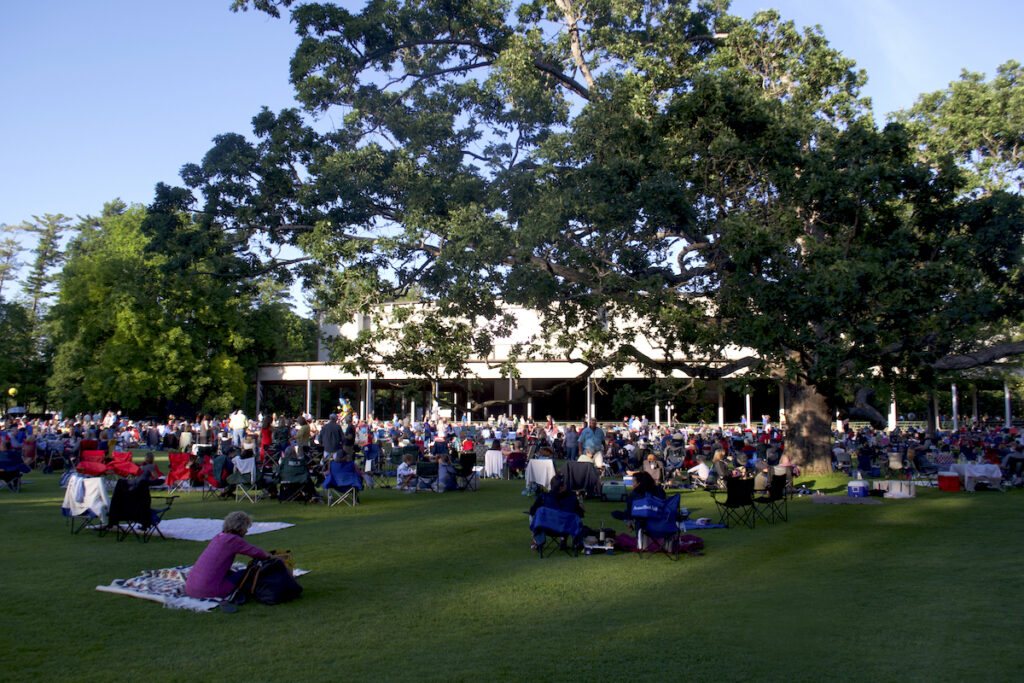People sit on blankets and lawn chairs on a grassy lawn under large trees, gathered near a covered stage or pavilion for an outdoor event.