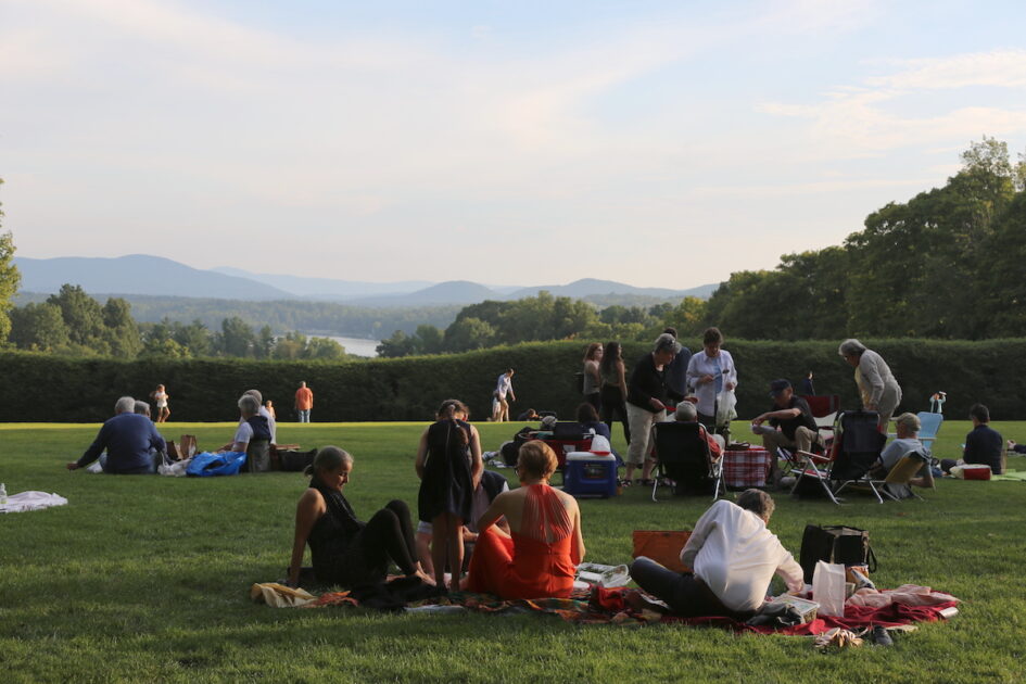 People gather on a grassy lawn for a picnic, seated on blankets and chairs, with trees and mountains visible in the background.