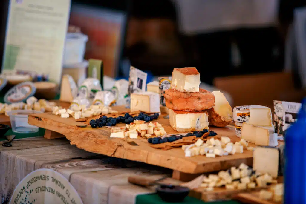 A wooden board displays various types of cheese, cut into cubes and wedges, with scattered blueberries and cheese labels on a table.