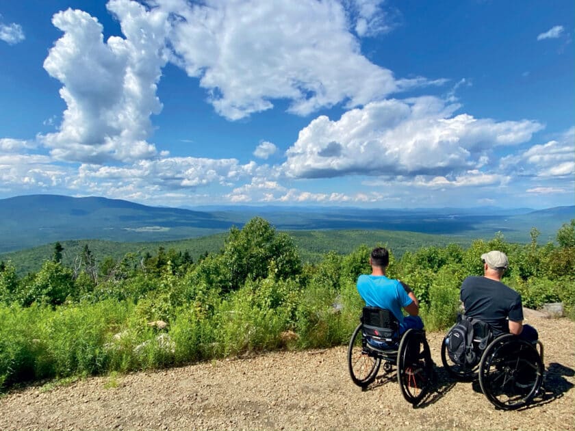 Accessible Travel in New England: Top Inclusive Adventures for a Barrier-Free Summer: Two people in wheelchairs sit on a dirt path overlooking a lush green valley with mountains and a cloudy sky in the background.