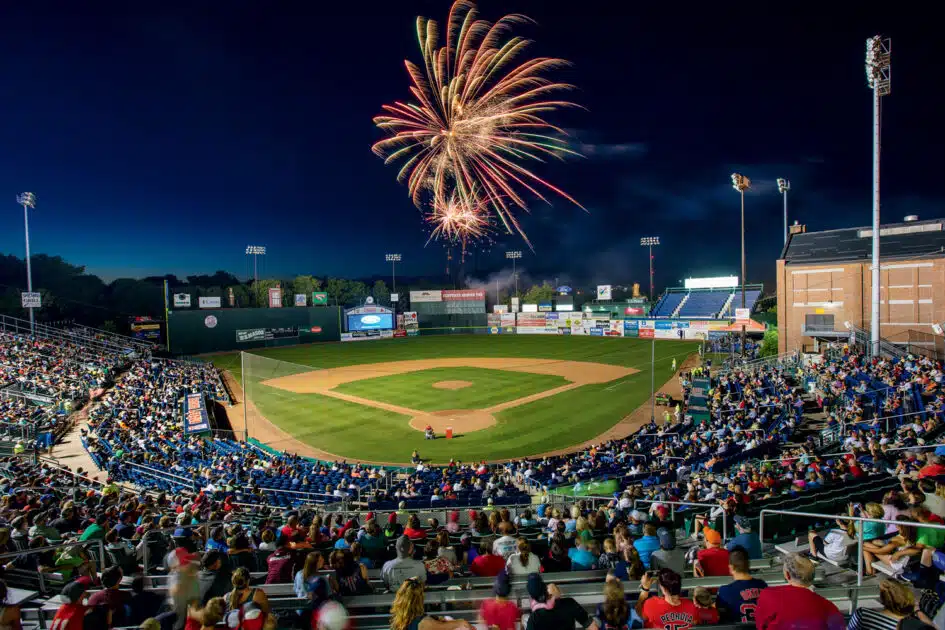 A baseball stadium filled with spectators at night, with fireworks lighting up the sky above the field.