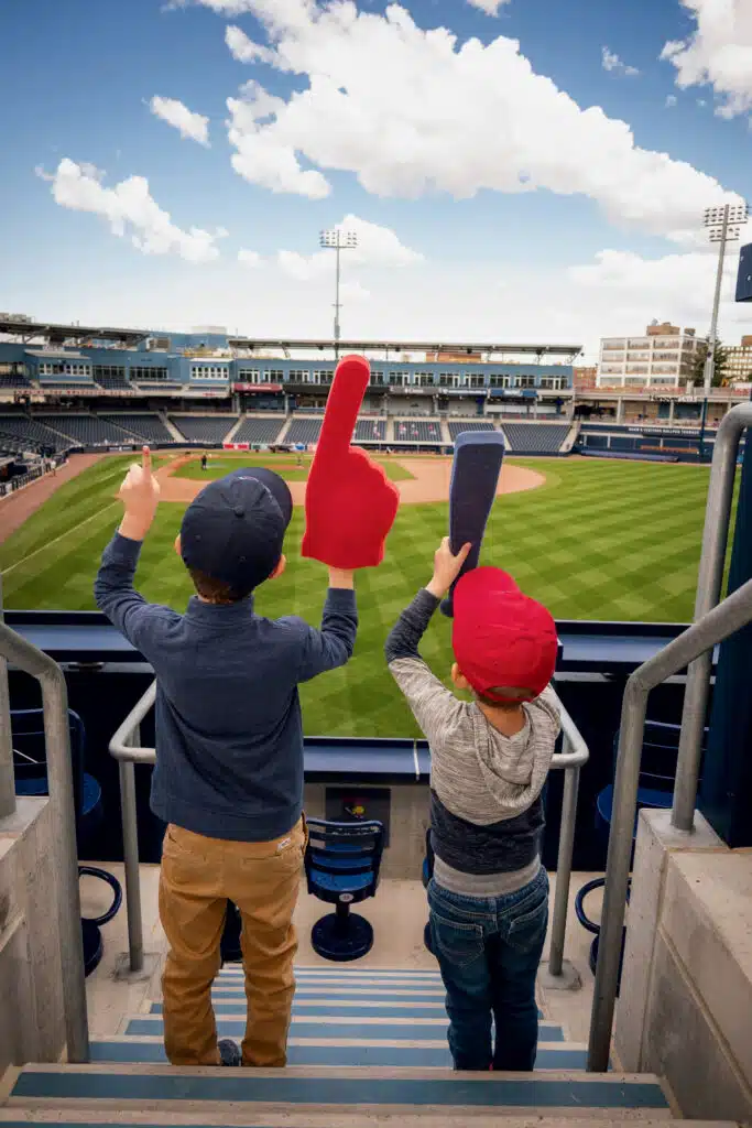 Minor League Baseball in New England: Big Fun, Small Parks | Two children wearing red caps stand in a stadium, holding foam fingers and facing an empty baseball field under a partly cloudy sky.