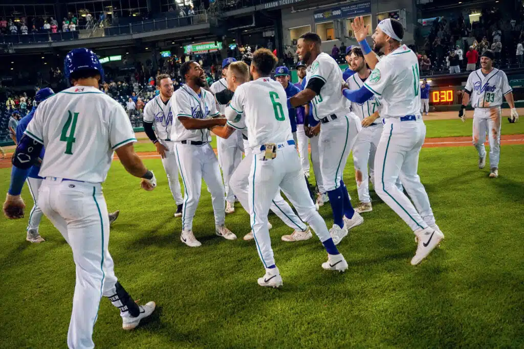 A group of baseball players in white uniforms celebrate together on the field at night, with stadium lights and spectators in the background.