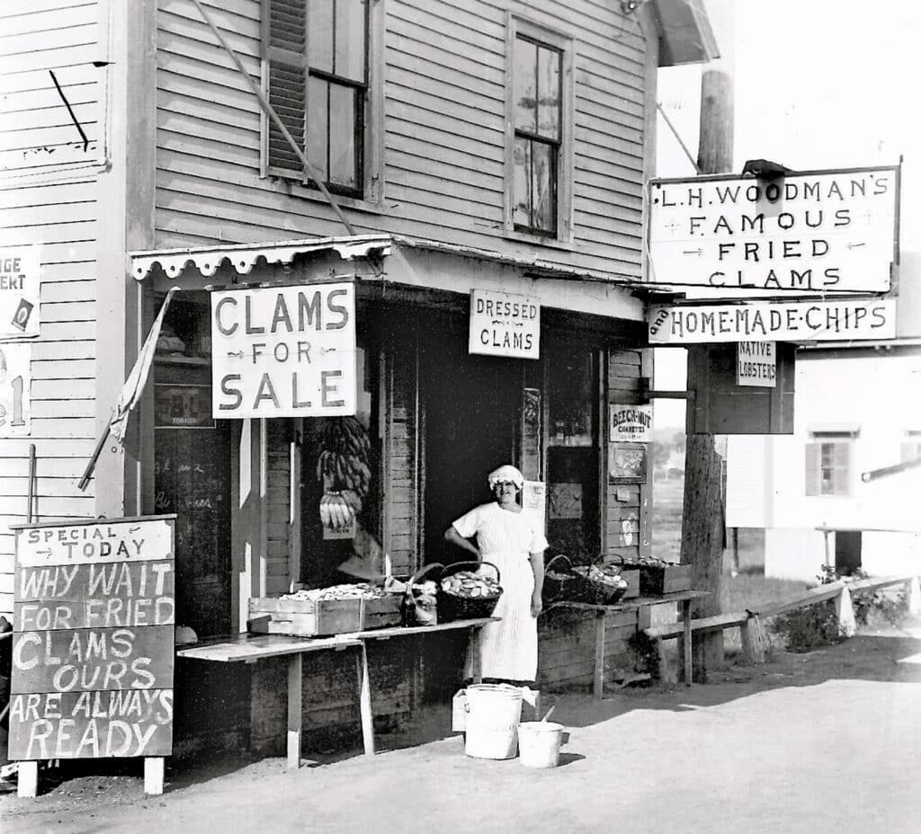 A person stands behind a stand selling clams and seafood outside a wooden building with multiple signs advertising clams and homemade chips.