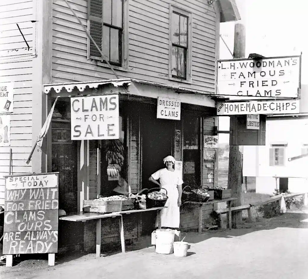A person stands behind a stand selling clams and seafood outside a wooden building with multiple signs advertising clams and homemade chips.