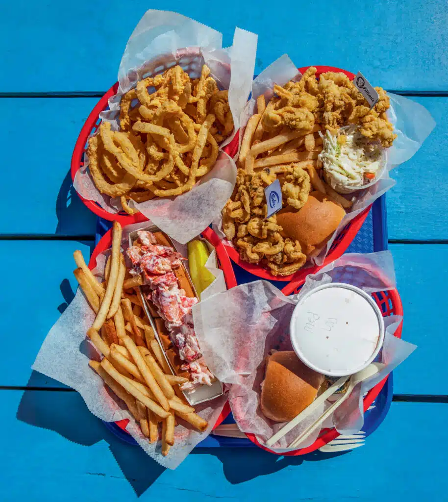 In Search of the Best Fried Clams in New England: A Coastal Road Trip. Overhead view of a tray with fried onion rings, fried seafood, coleslaw, a lobster roll with fries, a soft drink, and two bread rolls on a blue table.