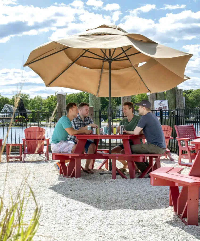 Four men sit at a round red picnic table under a large tan umbrella, talking and drinking canned beverages by a waterfront on a sunny day.