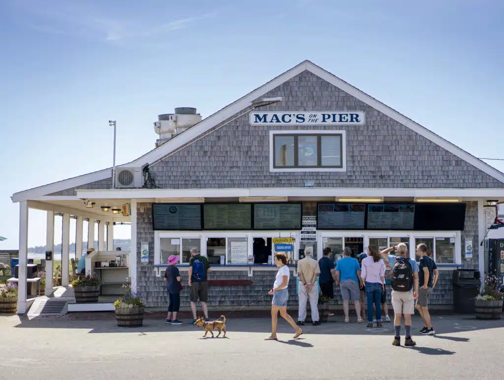 People stand in line at Mac’s on the Pier, a casual seafood restaurant with a gray shingle exterior under a clear blue sky.