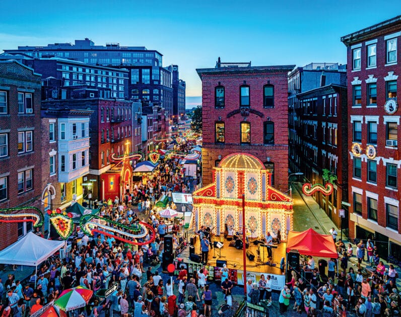 A large crowd gathers at an outdoor street festival with colorful lights, food stalls, and a decorated stage set between brick buildings at dusk.