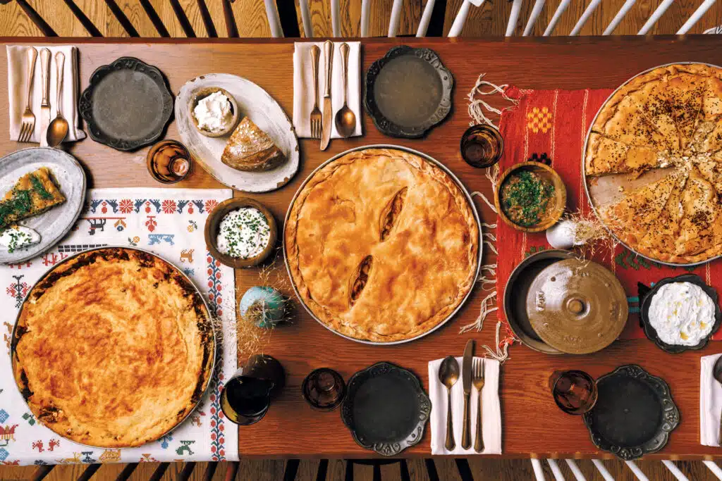 A table set for four with plates, utensils, glasses, various pies, side dishes, bread, and spreads on patterned tablecloths.