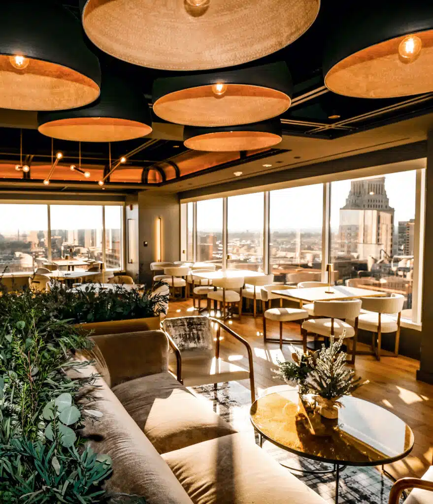 Best Things to Do in Connecticut This Summer 2025. Modern lounge with large windows, city views, beige seating, round tables, potted plants, and ceiling lights in daylight.