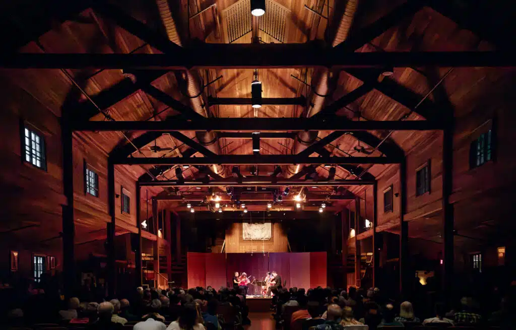A musical performance takes place on a stage in a large wooden hall, with an audience seated and watching under warm lighting.