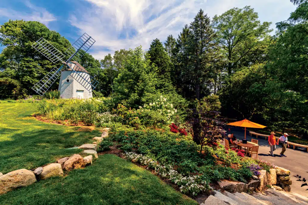 A scenic garden landscape with a windmill on the left, stone steps, lush greenery, and people gathered near a table with an orange umbrella on the right.