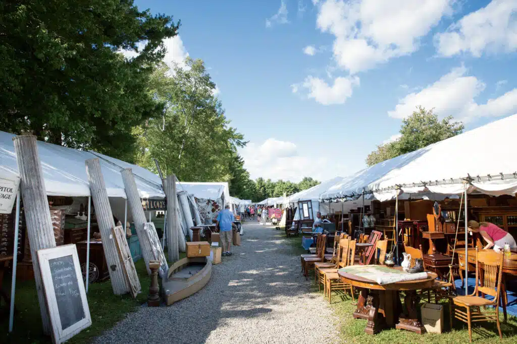 Outdoor flea market scene with white tents on both sides of a gravel path, displaying various furniture and antique items under a blue sky with scattered clouds.
