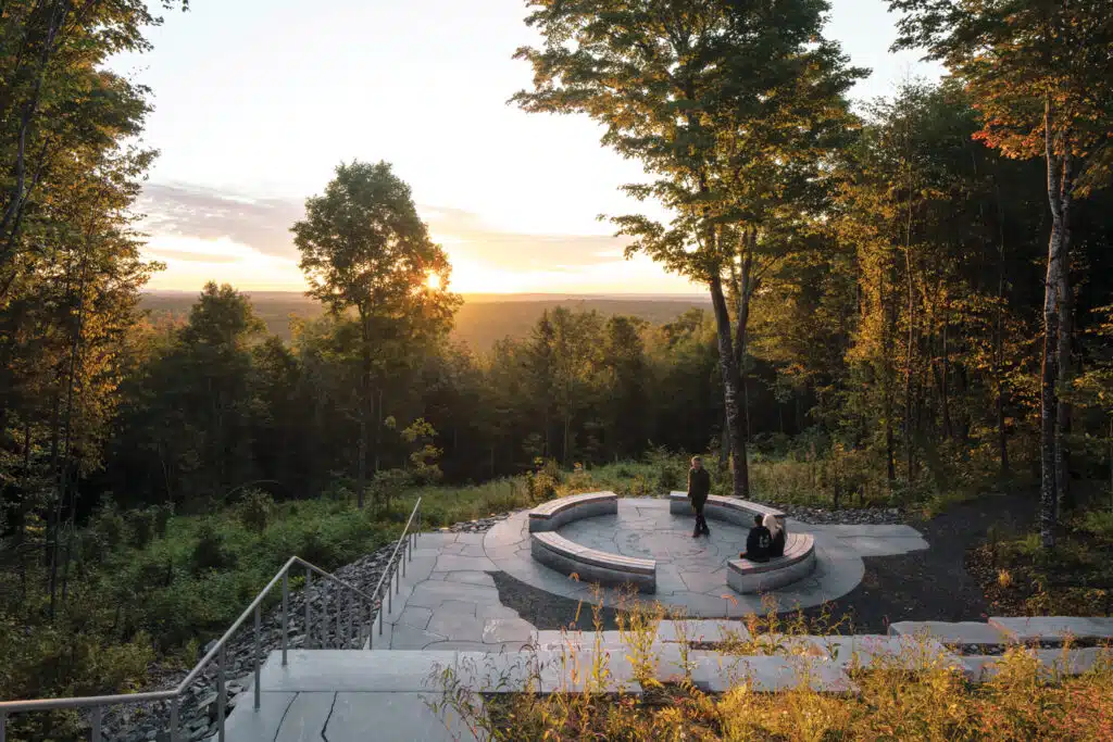 A circular stone seating area overlooks a forested landscape at sunset, with two people sitting on the benches surrounded by tall trees. 2025 Maine Travel Guide | Hotels, Dining & Attractions