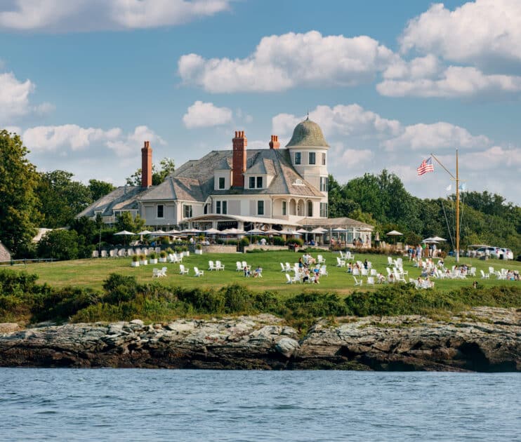Best Things to Do in Rhode Island This Summer. Large waterfront mansion with a round turret, manicured lawn, white outdoor chairs, people gathered, and an American flag, viewed from across the water under a partly cloudy sky.
